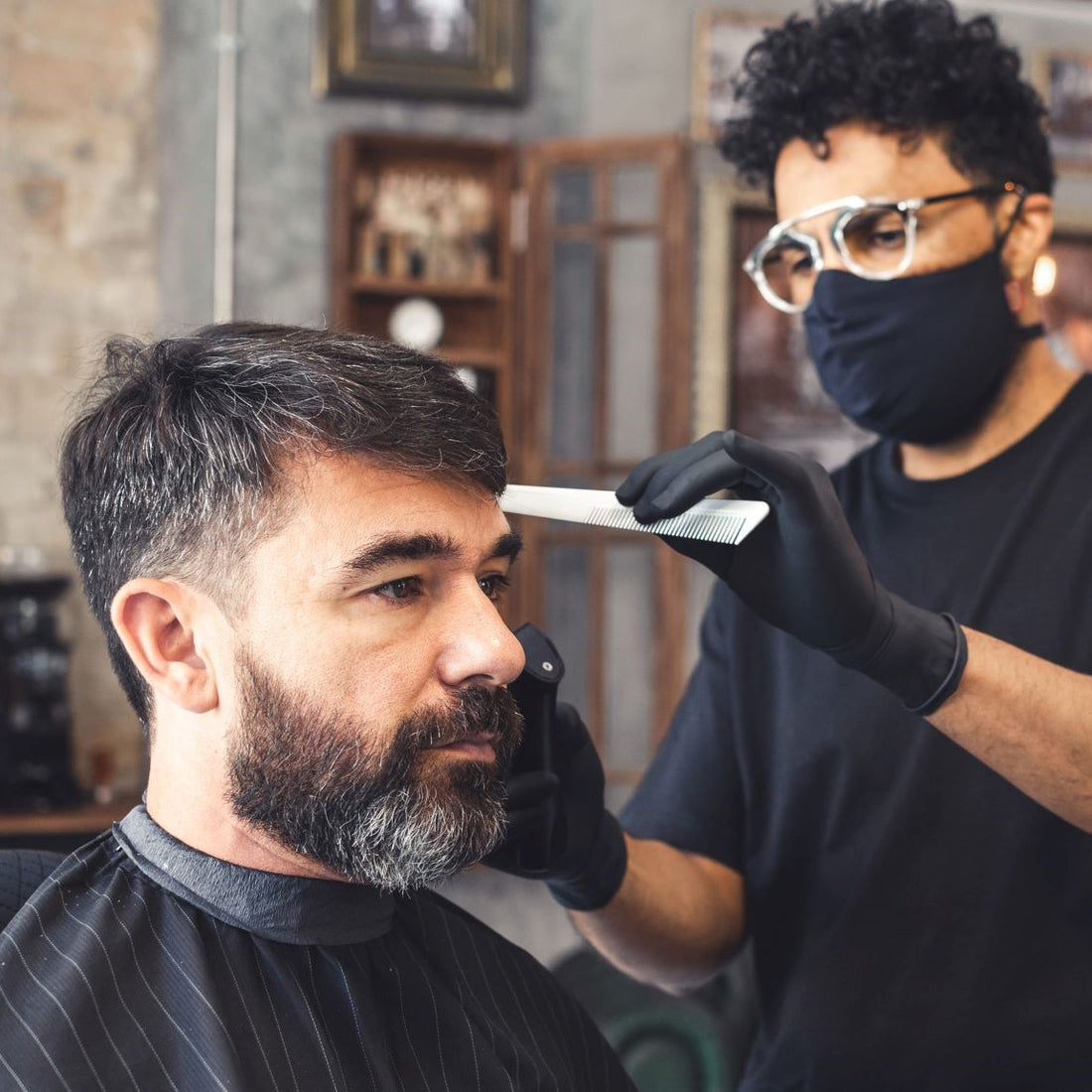 A male barber wearing a face mask cuts another man’s hair holding a comb and clippers.