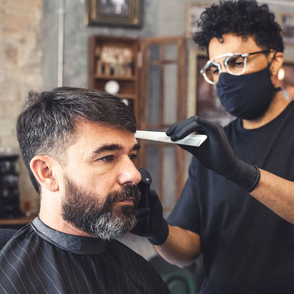 A male barber wearing a face mask cuts another man’s hair holding a comb and clippers.